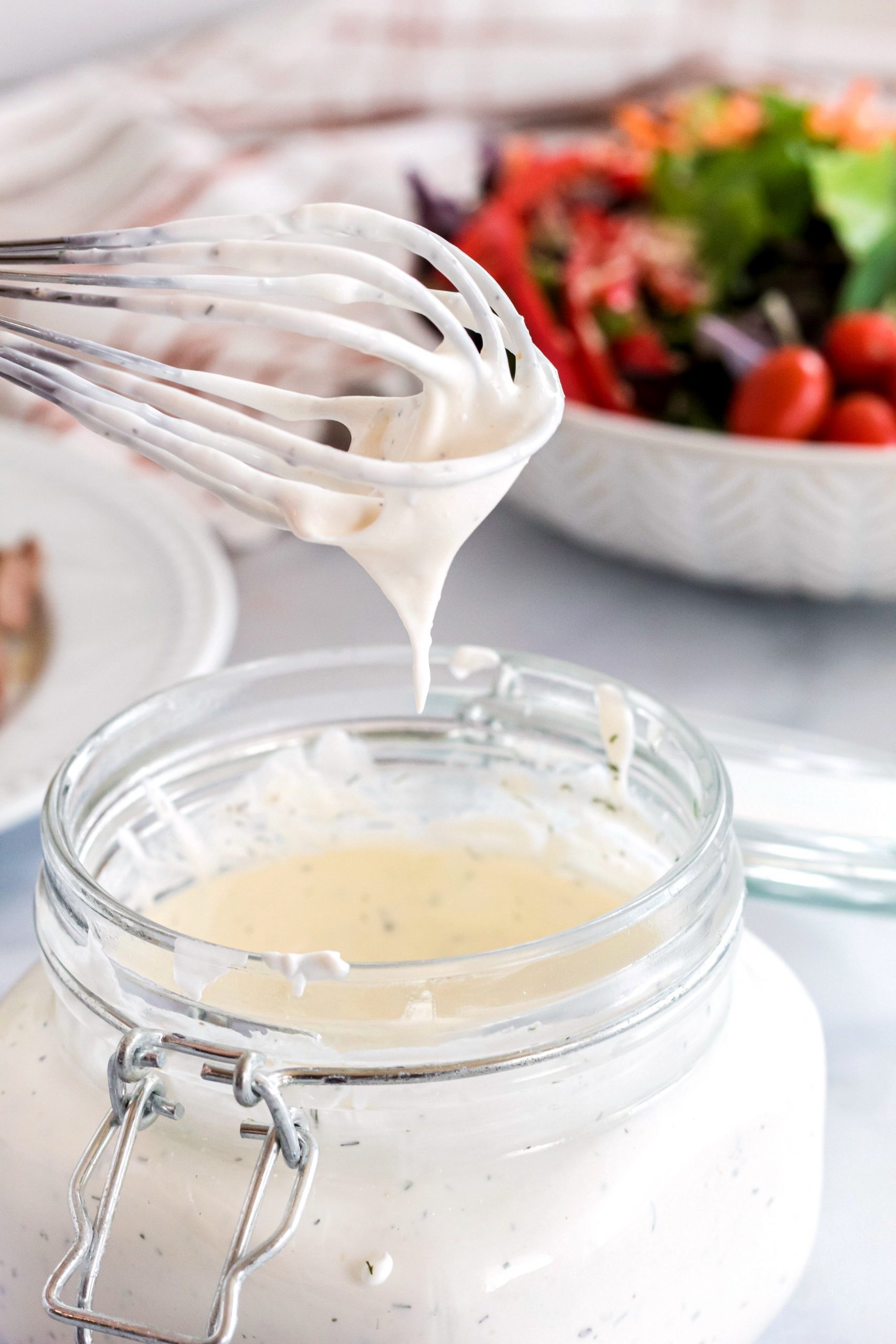 A jar of sour cream salad dressing that has been stirred with a wisk and a bowl of salad 