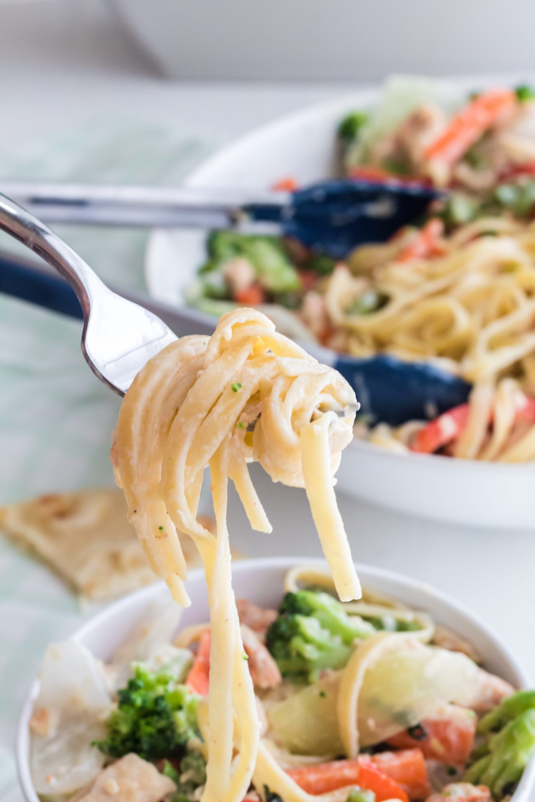 A fork full of fettuccine and a white bowl of fettccine with broccoli, carrots and red peppers