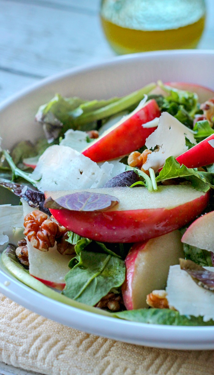 A combination of greens, apples, parmesan Cheese and walnuts in a white bowl