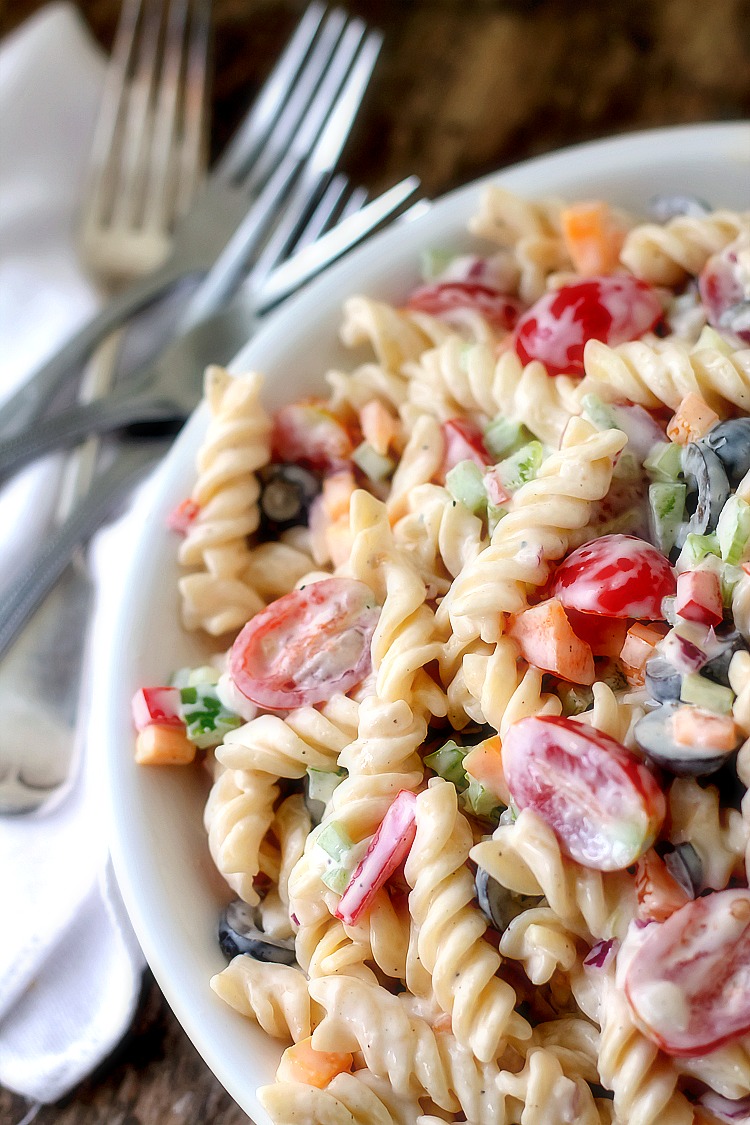 Pasta, tomatoes, carrots, peppers, olives topped with a decadent dressing in a white bowl