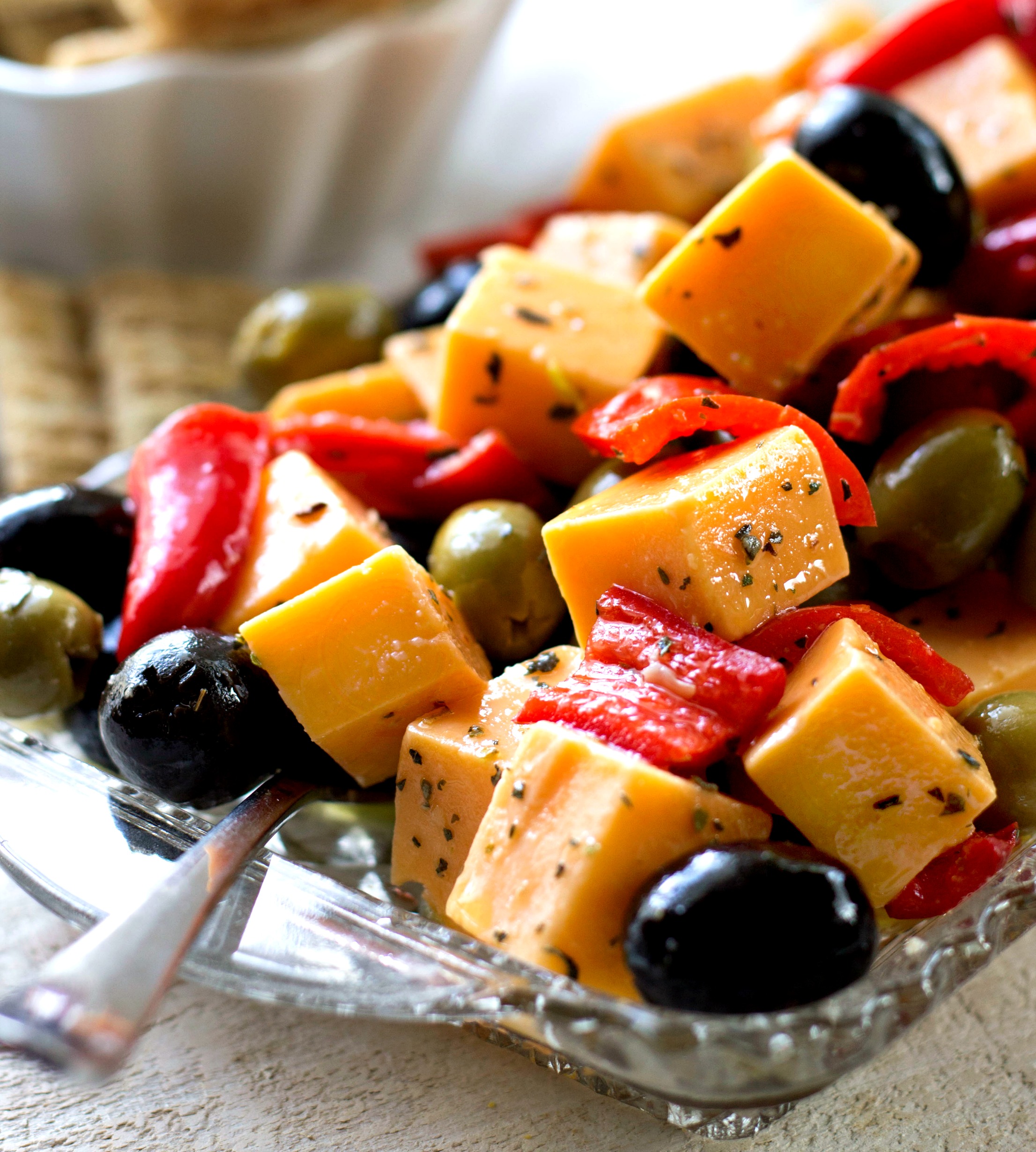 Cheese, olives, peppers that are topped with spices and seasoning in a pretty dish with crackers 