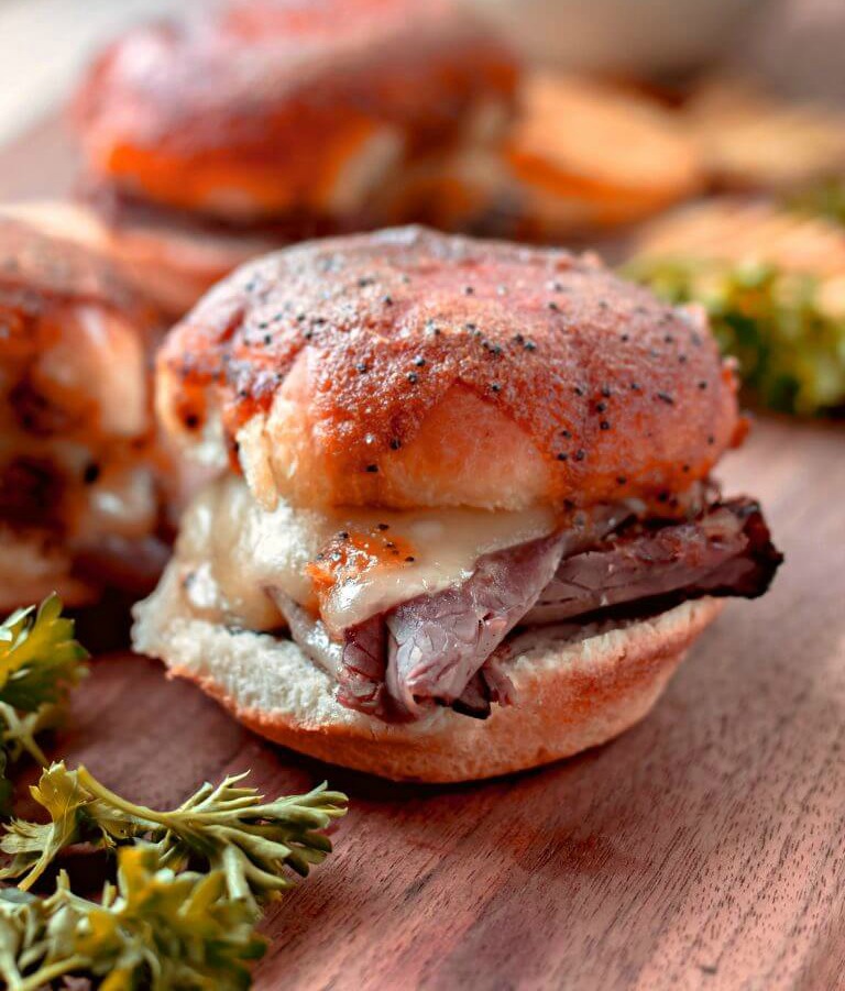 A few little roast beef and melted cheese sandwiches on mini buns on a wooden table