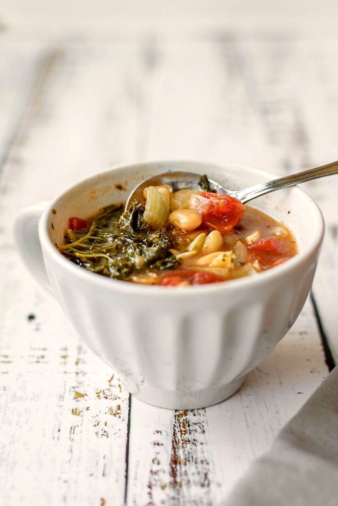White bowl with kale, beans and lots of veggies in a delicious soup on a white table