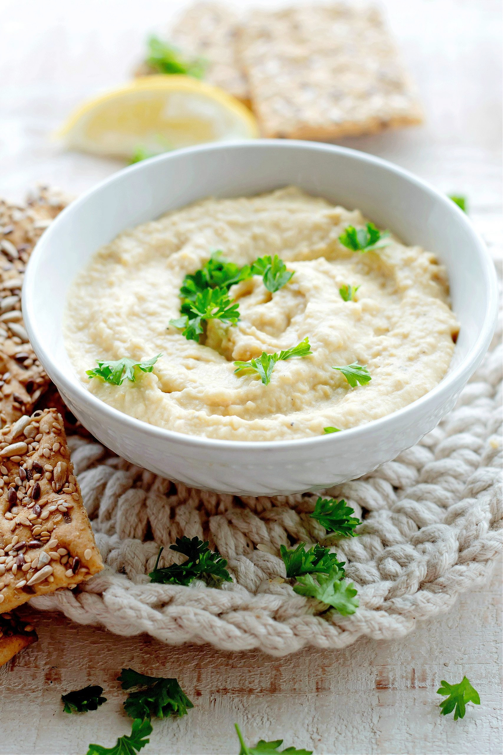 Creamy hummus topped with parsley in a white bowl and surrounded by crackers